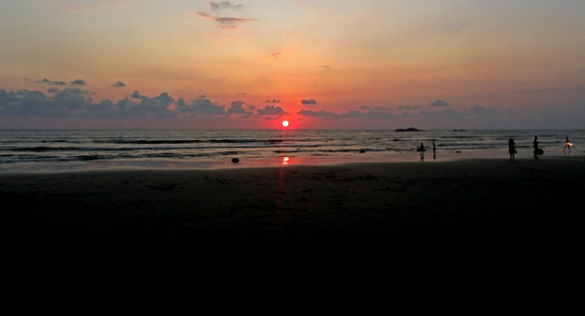 A red sun sets on the horizon of the ocean. In the foreground, the silhouettes of people dot the shore.